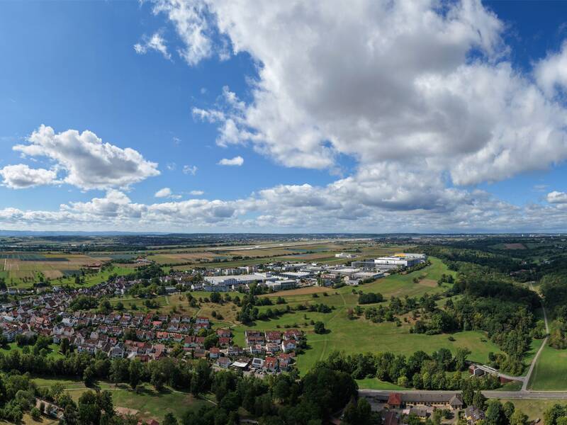 Das Bild zeigt eine weitläufige Landschaft mit einer Mischung aus Feldern, Waldgebieten und einer Siedlung unter einem blauen Himmel mit vielen Wolken.