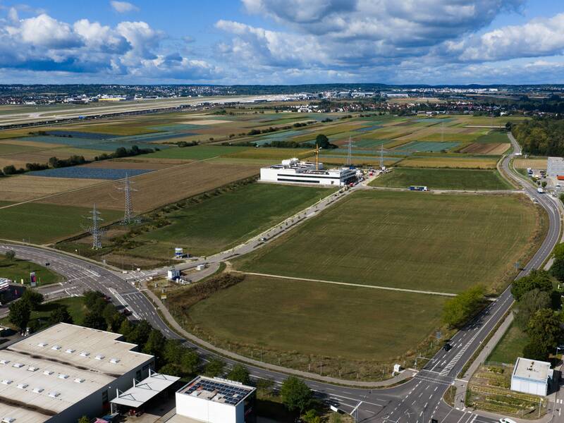 Luftaufnahme einer ländlichen Landschaft mit Feldern, einer Flughafenlandebahn im Hintergrund und mehreren Straßen und Gebäuden im Vordergrund unter einem teils bewölkten Himmel.