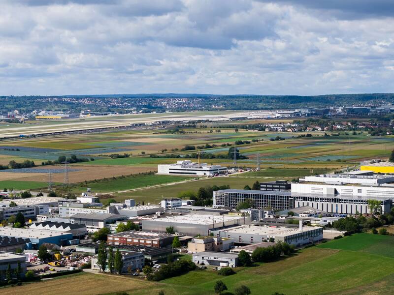 Luftaufnahme eines weitläufigen Gewerbegebiets mit angrenzenden Feldern und einem dahinter liegenden Flugplatz.