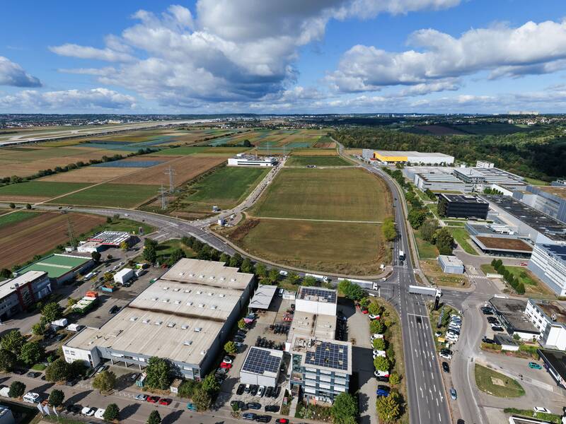 Luftaufnahme einer weitläufigen Landschaft mit landwirtschaftlichen Feldern, Straßen und mehreren Gebäudekomplexen unter einem teilweise bewölkten Himmel.